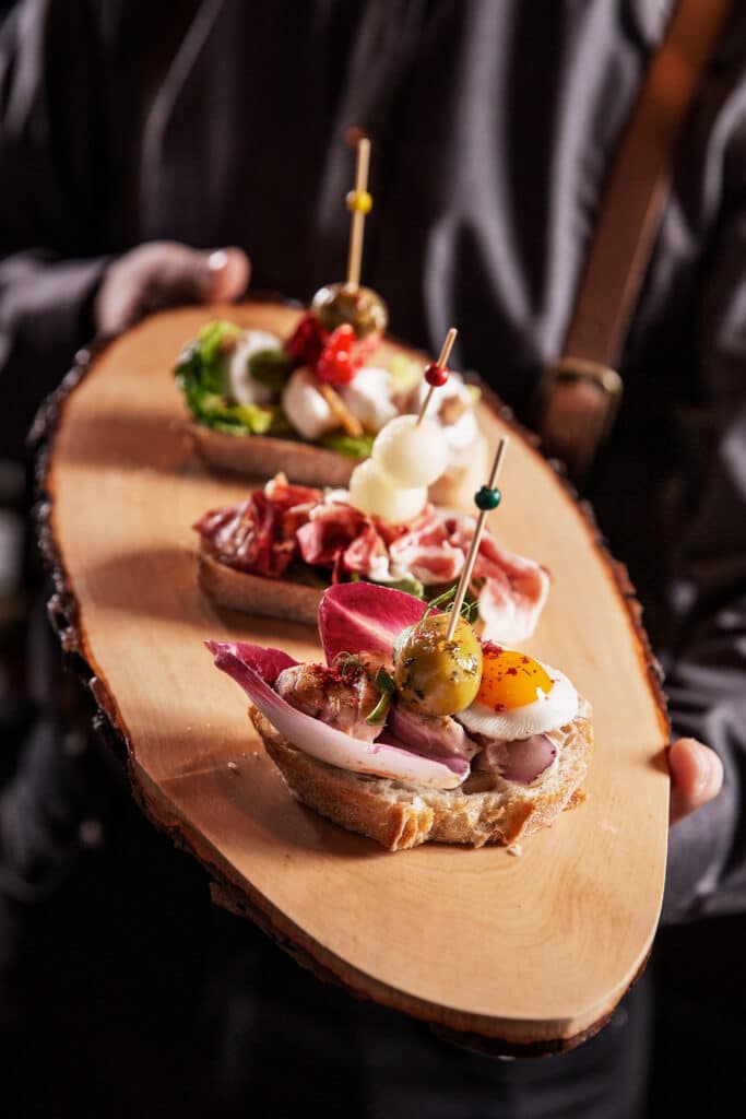Een houten plateau wordt gepresenteerd met daarop drie smakelijk opgemaakte bruschetta's, elk belegd met diverse luxe ingrediënten zoals vleeswaren, groentes en garnituren, vastgezet met kleurrijke prikkers. De focus ligt op de combinatie van kleuren en texturen, passend bij een gastronomische ervaring.