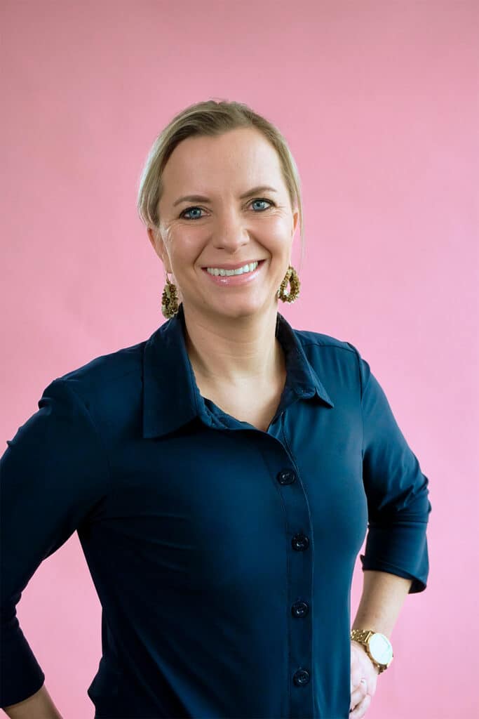 Marleen op een roze achtergrond met een donkerblauwe blouse en de handen in de zij