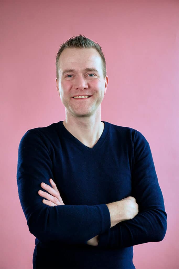 Sjoerd op een roze achtergrond met een donkerblauwe trui en handen over elkaar