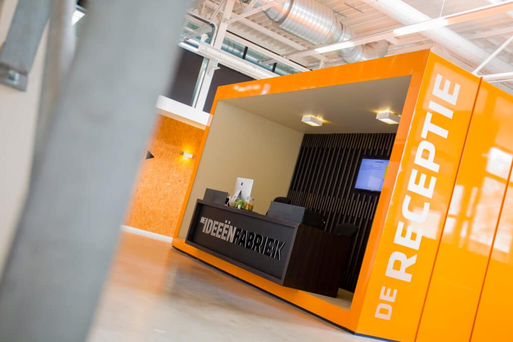 Een moderne receptie met felle oranje panelen aan de buitenkant. Het woord "RECEPTIE" is zichtbaar op de zijkant. Binnen staat een houten balie met computers. Het plafond heeft een industrieel uiterlijk met zichtbare luchtkanalen.