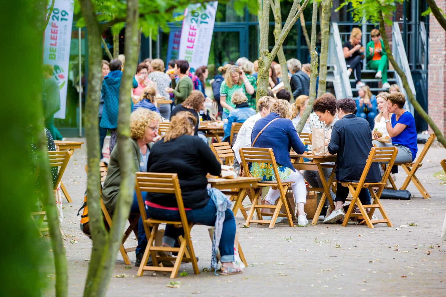 Mensen zitten buiten aan houten tafels en stoelen tijdens een evenement. Er zijn bomen op de voorgrond en vlaggen met "Beleef 2019" op de achtergrond.