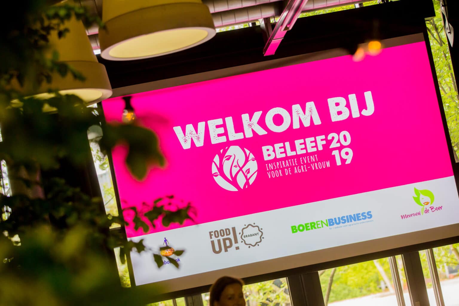 Scherm met roze achtergrond dat het "Beleef 2019" inspiratie-evenement voor agri-vrouwen verwelkomt. Vooraan zichtbare groene planten en zichtbare logos onderaan.
