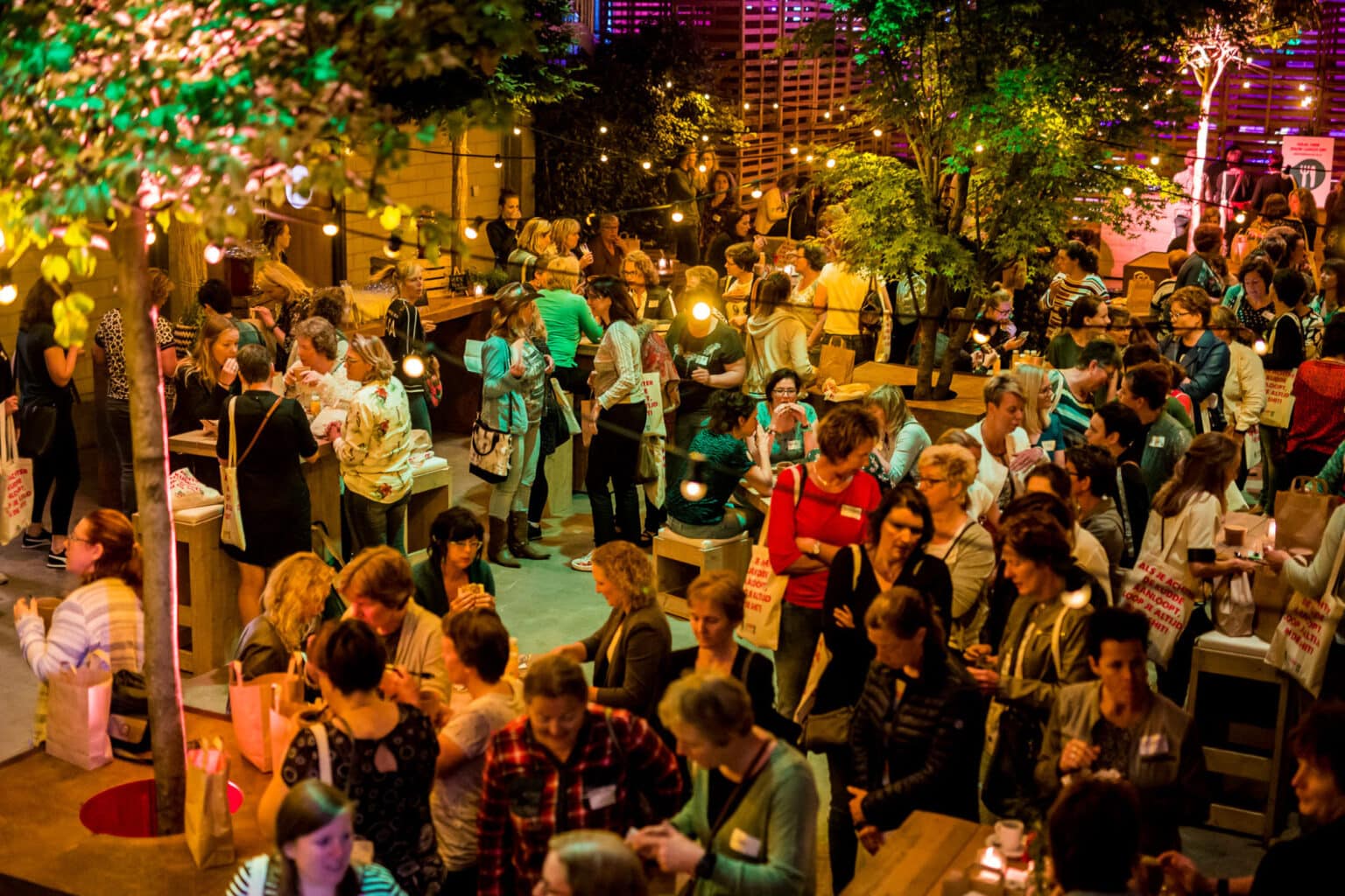 Drukke en sfeervol verlichte netwerkevenement in een overdekte ruimte met veel mensen die in groepjes praten, eten en drinken. Er hangen lichtslingers en er is veel groen, wat de setting gezellig en uitnodigend maakt.