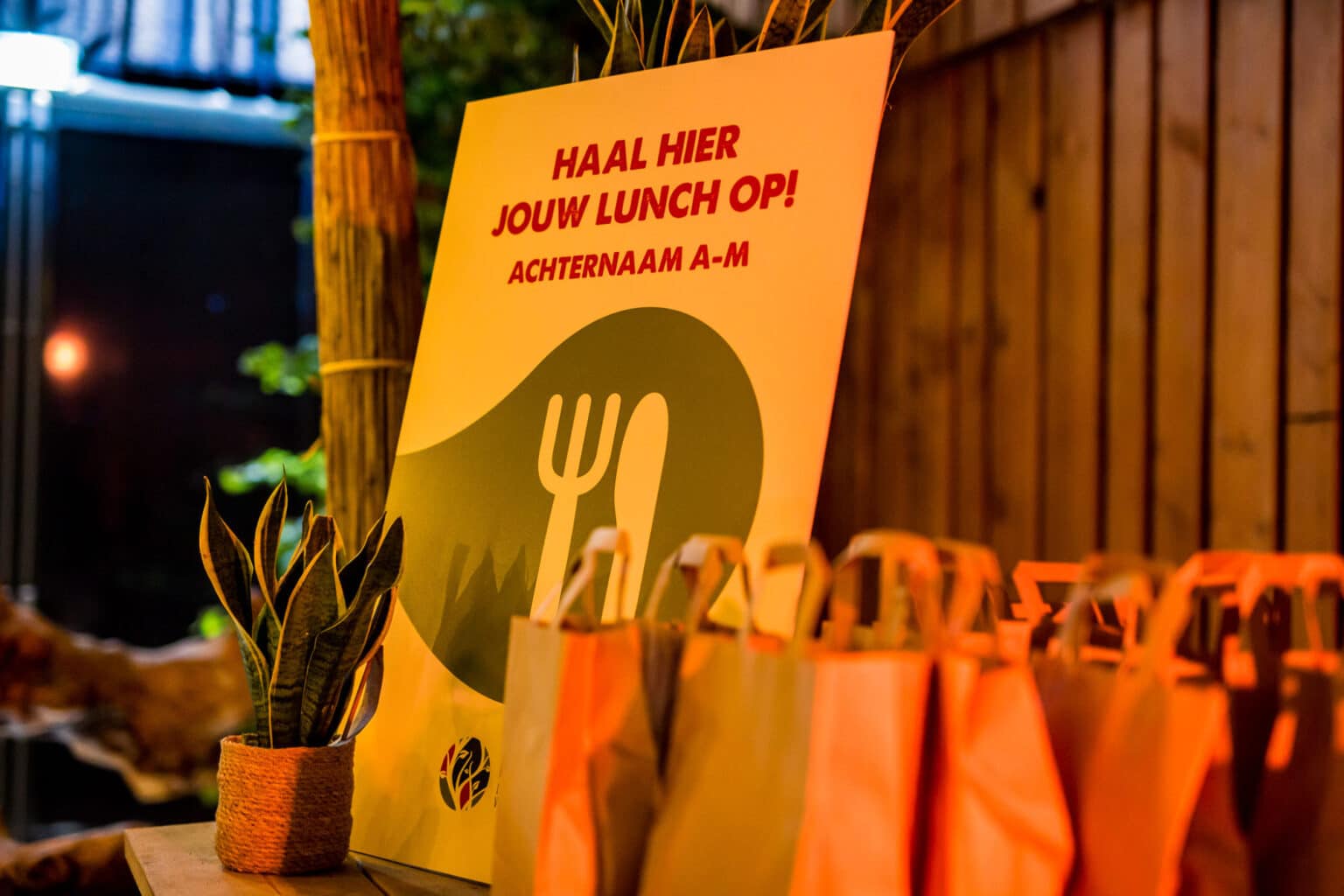 Een bord met de tekst “Haal hier jouw lunch op! Achternaam A-M” staat op een tafel. Ernaast staan papieren lunchzakken en een kleine kamerplant in een pot. De ruimte is warm verlicht, en op de achtergrond is een houten wand te zien.