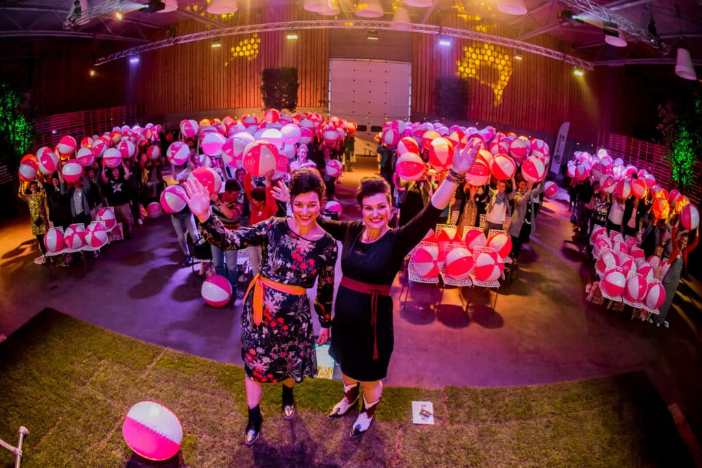 Twee vrouwen staan voor een feestelijke menigte die roze en witte strandballen vasthouden. De zaal is sfeervol verlicht met gekleurde lichten, en er hangen decoraties aan het plafond. De vrouwen glimlachen en zwaaien naar de camera. In de achtergrond is de menigte vrolijk en energiek.
