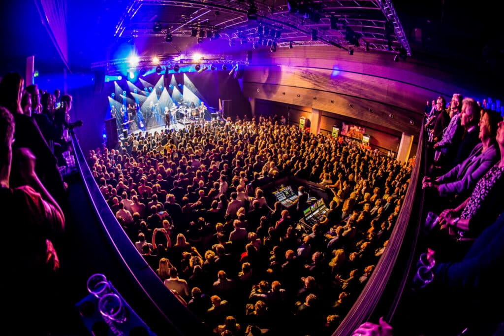 Een volle zaal in Markant Uden, er wordt een concert gegeven. Er staan mensen op de vloer en bovenaan op een soort balkon.