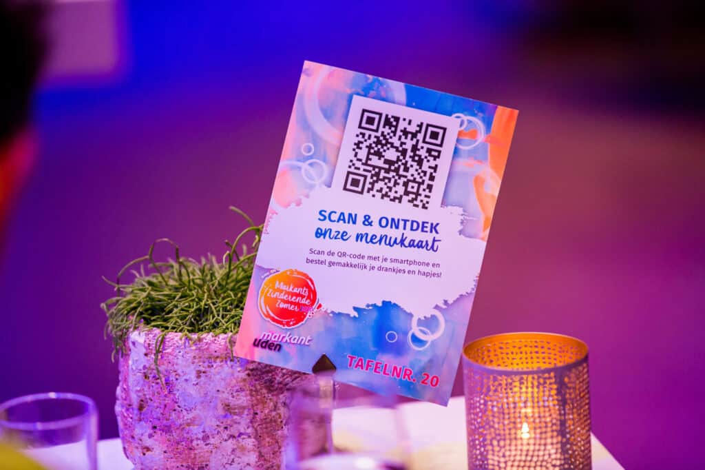 Een kaart op een tafel met een kleurrijke achtergrond en een QR-code. De tekst op de kaart nodigt uit om de menukaart te scannen met een smartphone voor het bestellen van drankjes en hapjes. Er is decoratie met een kaars en een plant te zien op de tafel. Het tafelnummer is "20".