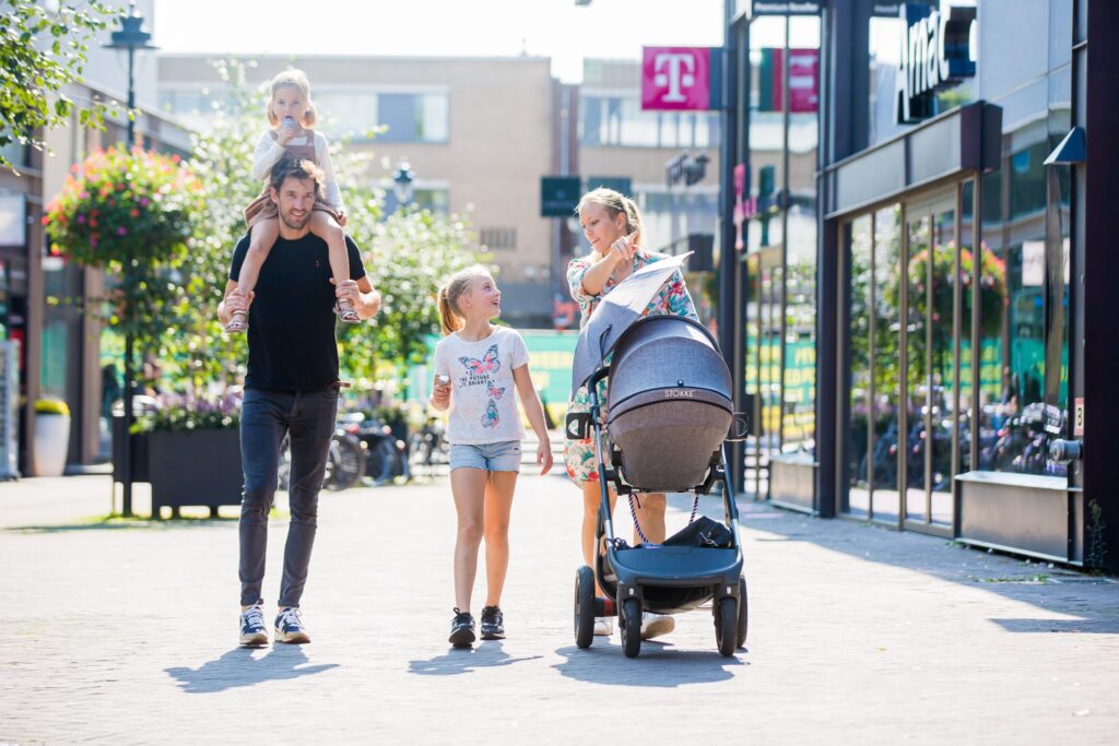 Een gezin loopt samen door een winkelstraat op een zonnige dag. Een vader draagt een kind op zijn schouders. Naast hen loopt een oudere dochter, en een moeder duwt een kinderwagen. De straat is levendig, omringd door winkels en beplante bloembakken.