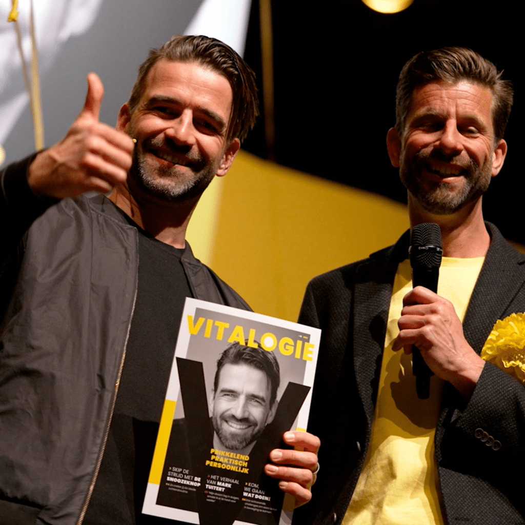 Twee mannen op een podium, één houdt een tijdschrift "Vitalogie" vast en steekt een duim op.