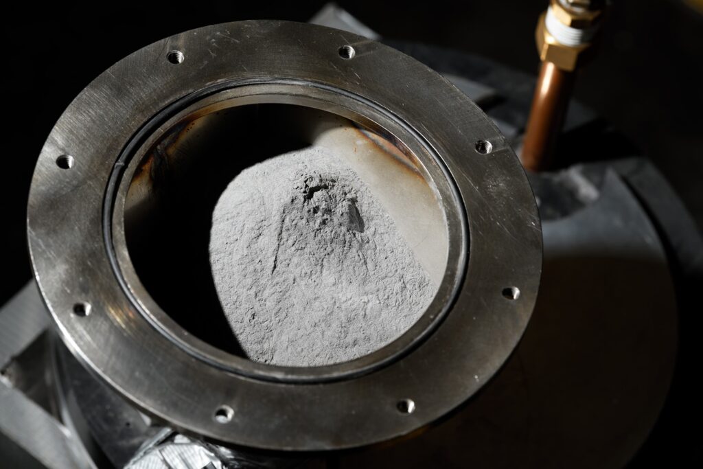 Een metalen vat gevuld met een grijs, poederachtig materiaal, dat centraal in beeld is geplaatst. Het vat heeft een robuuste metalen rand met bouten, en er is een koperen buis zichtbaar aan de zijkant. Het poeder bevindt zich in een donkere, industriële omgeving.