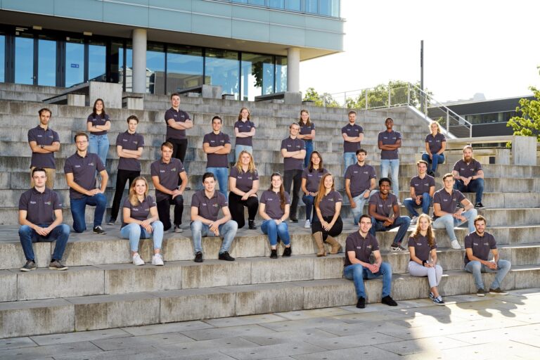 Een groep mensen zit en staat op de trappen voor een gebouw. Ze dragen allemaal bijpassende donkergrijze T-shirts met logo's. Het lijkt een formele groepsfoto te zijn, mogelijk van een team of klas. Op de achtergrond is een modern gebouw met glazen ramen zichtbaar.
