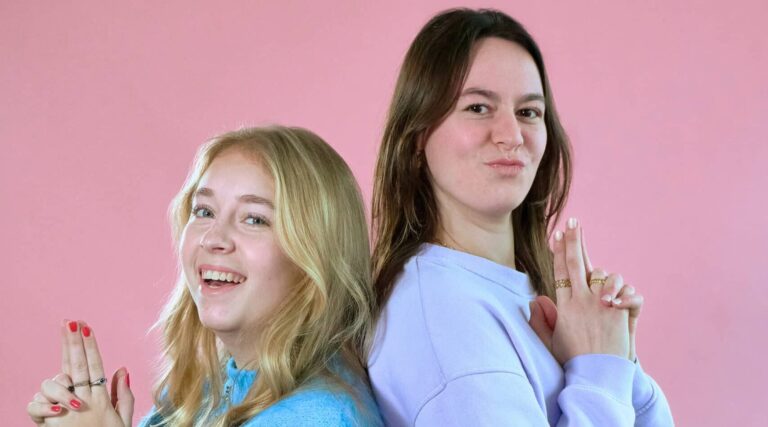 2 vrouwen op een roze achtergrond 1 met een blauwe trui en 1 met een paarse trui. Blond haar en donker haar.