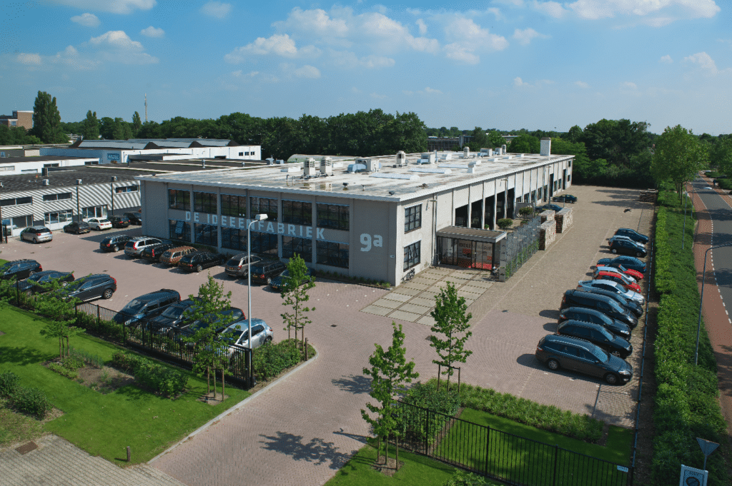 Een luchtfoto van een groot industrieel gebouw met de naam "De Ideeënfabriek" op de voorkant. Het gebouw heeft een ruim parkeerplaats.