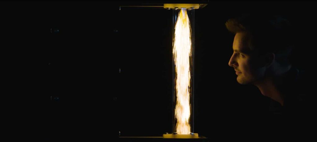 Een man kijkt naar een verticale kolom van vonken of lichtstralen in een donkere omgeving. De lichteffecten creëren een sterk contrast tegen de zwarte achtergrond. Het gezicht van de man is subtiel verlicht door het lichtfenomeen.