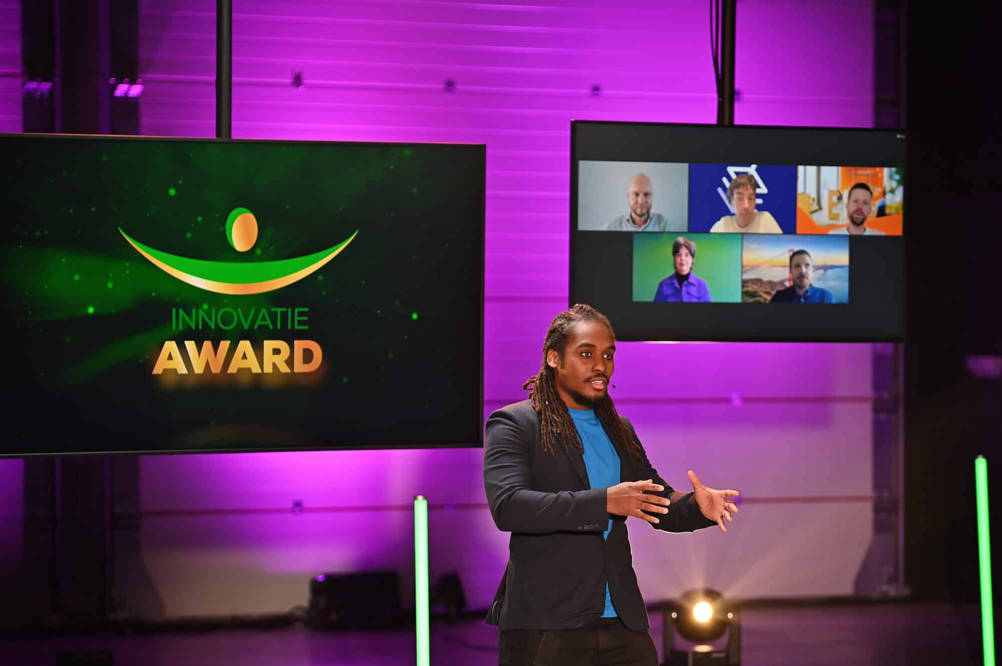 Een presentator spreekt op een podium tijdens een evenement met de naam "Innovatie Award". Op de achtergrond zijn grote schermen te zien; op één scherm staat de tekst "Innovatie Award" met een grafisch logo, terwijl op het andere verschillende mensen in een videoconferentie zichtbaar zijn. De verlichting in de ruimte is voornamelijk paars en groen.