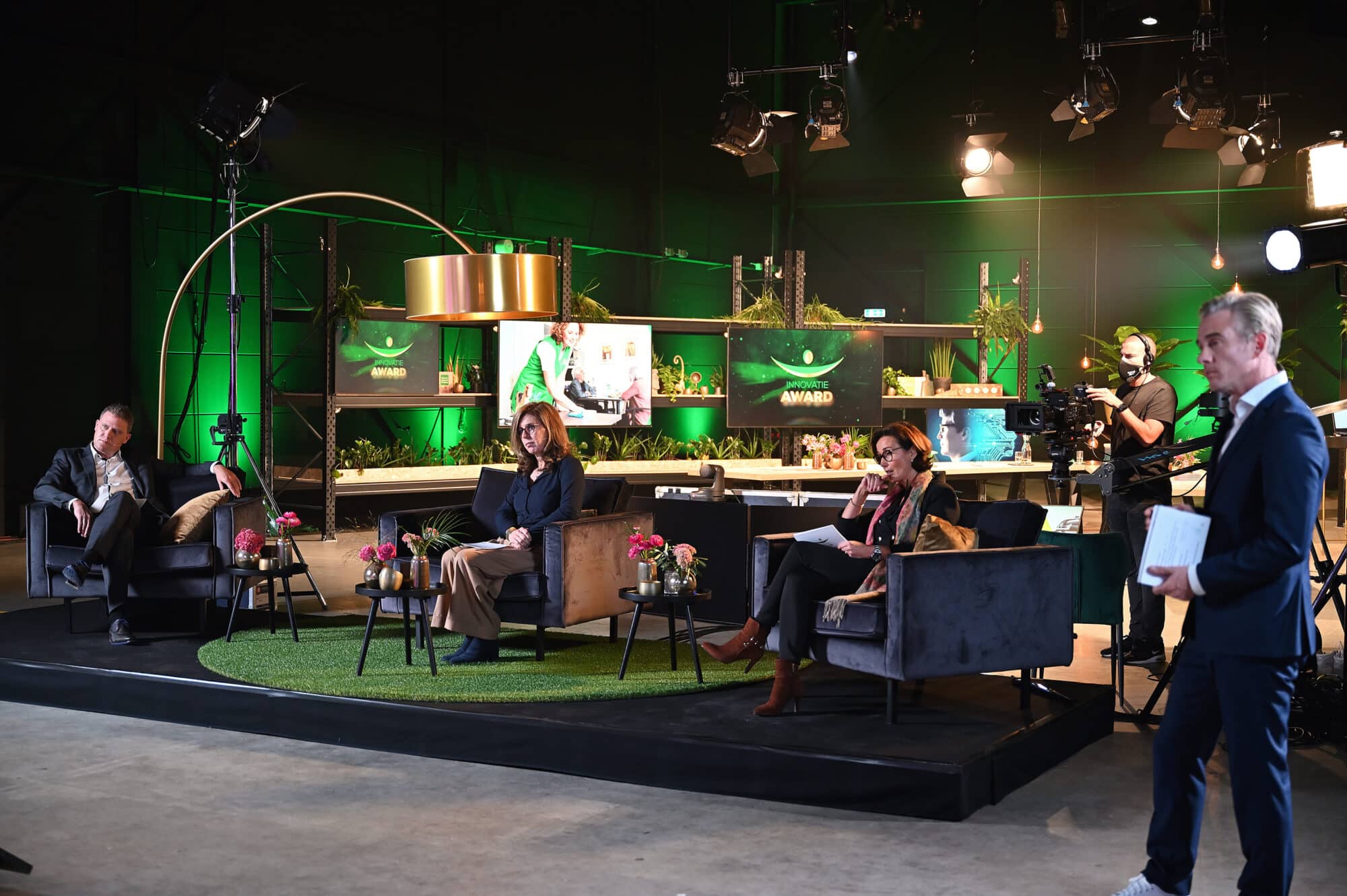 Een televisiestudio met drie mensen die op stoelen zitten te midden van decor met planten en schermen met het "Innovatie Award" logo. Een cameraman staat klaar met een camera, terwijl een presentator op de voorgrond staat met papieren in de hand. De sfeer is professioneel en de verlichting is helder.