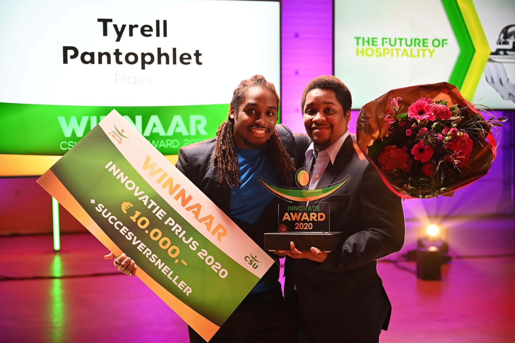 Twee personen poseren blij met een cheque en een trofee voor de Innovatie Prijs 2020. Ze houden een groot bord vast waarop "Winnaar Innovatie Prijs 2020" en "€10.000" staat. Er is ook een bos bloemen zichtbaar. Op de achtergrond zijn schermen te zien met teksten zoals "The Future of Hospitality".