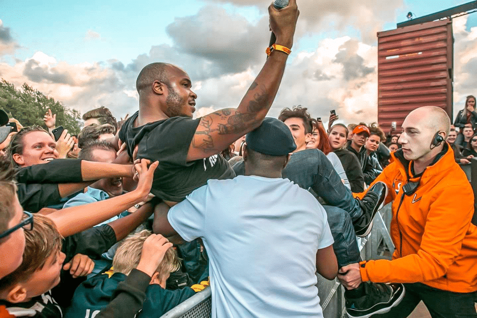 Een artiest met een microfoon wordt door een enthousiaste menigte op handen gedragen tijdens een festival. De beveiliging helpt een crowdsurfer over de dranghekken terwijl de sfeer energiek en uitgelaten is. Op de achtergrond kijken festivalgangers vol enthousiasme toe onder een bewolkte hemel.