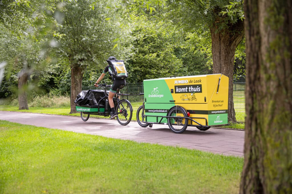 Een man fietst met een grote bakfiets langs een groen park. De bakfiets is uitgerust met een aanhangwagen waarop reclame staat van een supermarkt. De omgeving is omgeven door bomen en gras.