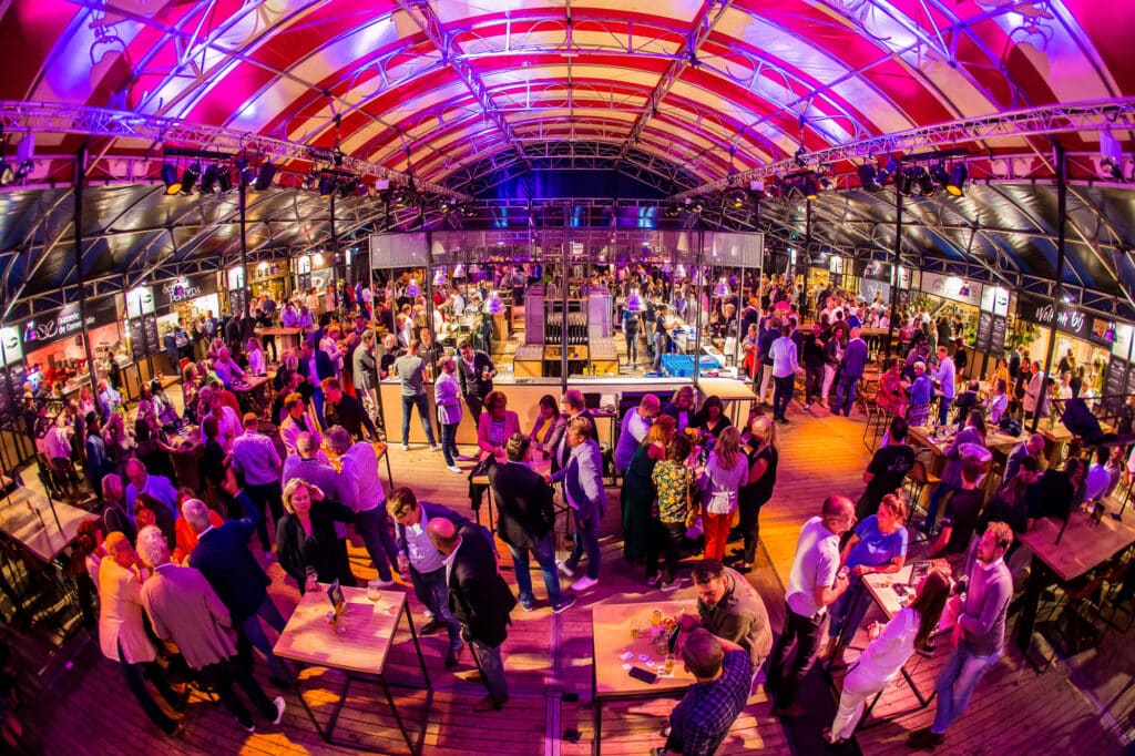 Drukke evenementenlocatie met mensen die netwerken onder kleurrijke verlichting in een grote overdekte ruimte. Een grote tent met rood-witte strepen.