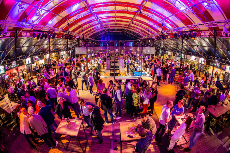 Drukke evenementenlocatie met mensen die netwerken onder kleurrijke verlichting in een grote overdekte ruimte. Een grote tent met rood-witte strepen.