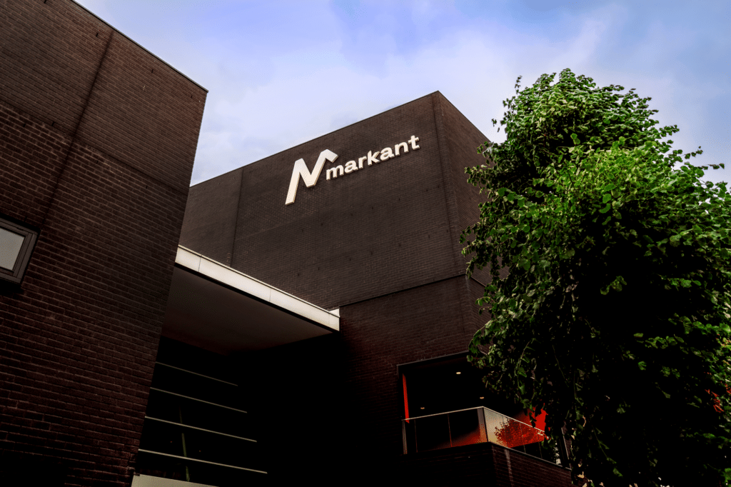 Een modern gebouw met donkere baksteen, waarop het logo "Markant" prominent is weergegeven. Er is een groene boom zichtbaar naast het gebouw, en de lucht is helder met een paar wolken. Het ontwerp van het gebouw heeft een strakke en eigentijdse uitstraling.