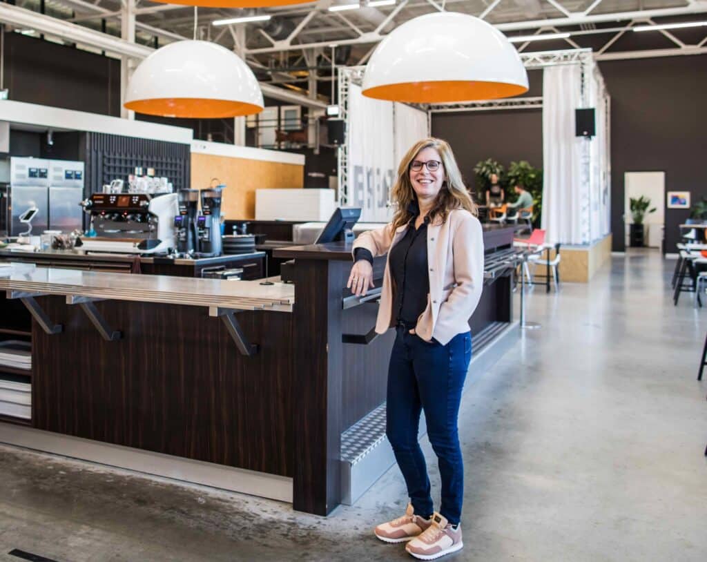 Een vrouw staat glimlachend naast een moderne, houten koffiebar in een ruim en open interieur. Boven de bar hangen grote, ronde lampen. In de achtergrond zijn industriële elementen en planten te zien, wat de ruimte een eigentijdse en uitnodigende sfeer geeft. Ze draagt casual kleding en sneakers.