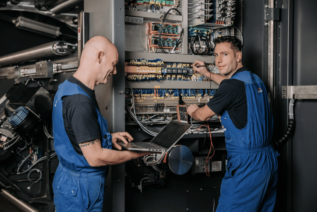 Twee technici in blauwe overalls zijn aan het werk bij een grote schakelkast vol met draden en elektronische componenten. De ene gebruikt een laptop en lacht, terwijl de andere bezig is met bekabeling. De omgeving heeft een industriële uitstraling.