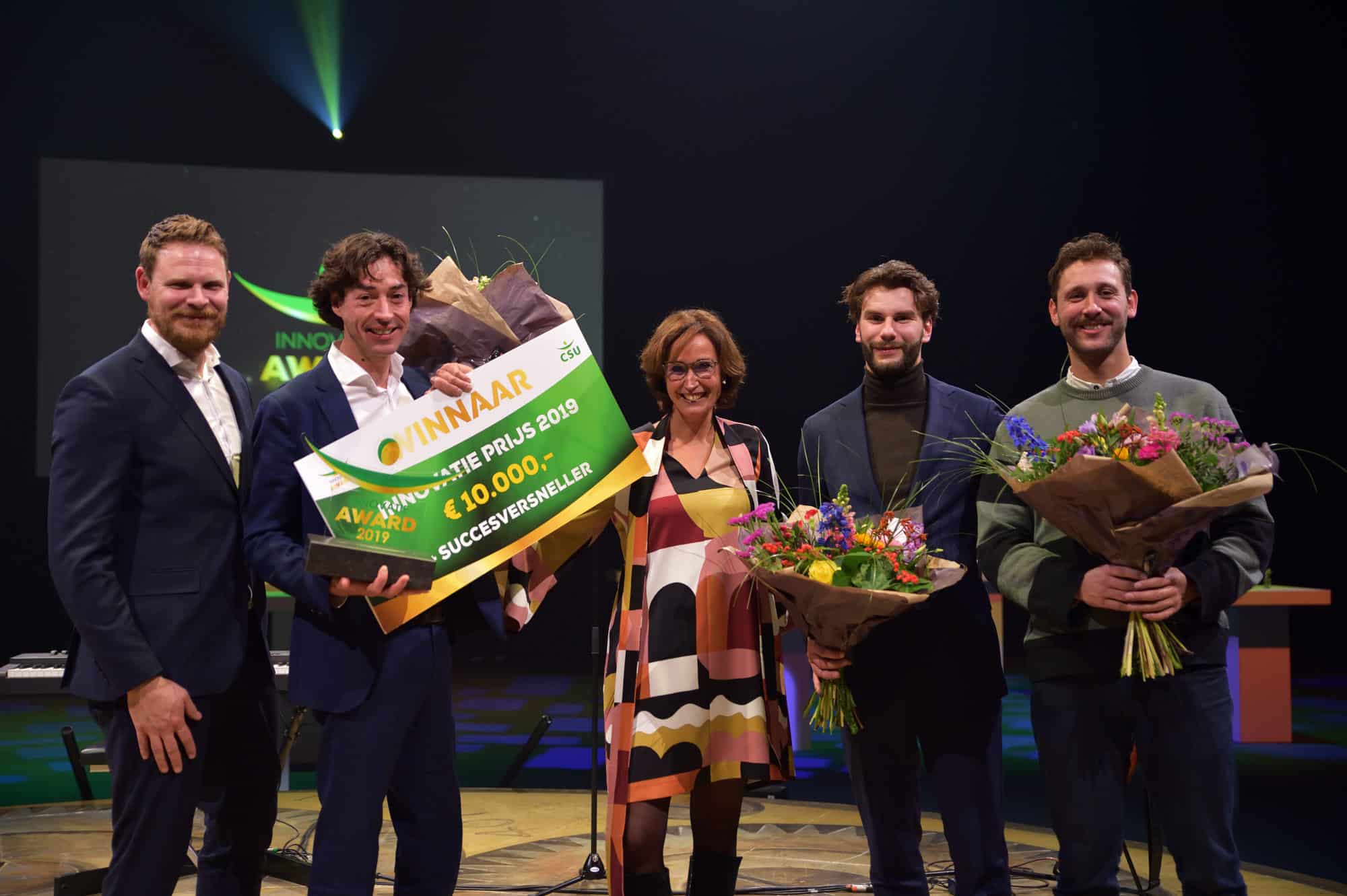 Een groep mensen poseert op een podium tijdens de Innovatie Award 2019. Ze houden grote boeketten bloemen vast en een groot cheque-bord waarop "Winnaar Innovatie Prijs 2019" en "€10.000" staat. Op de achtergrond is een scherm met het logo van de Innovatie Award te zien.