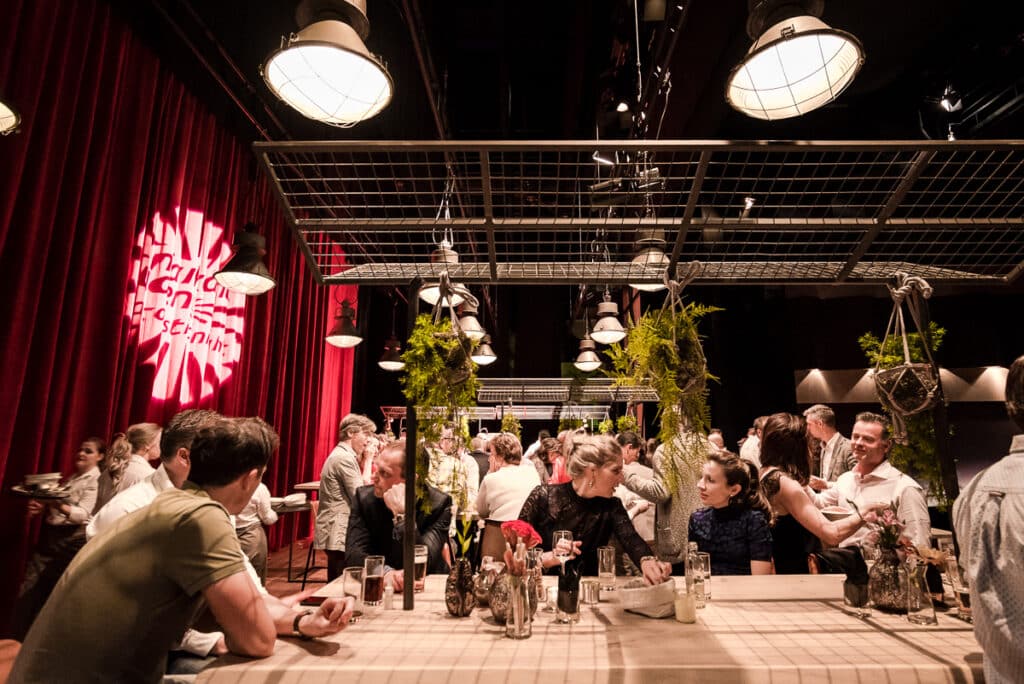 Een levendige bijeenkomst in een sfeervolle ruimte met rode gordijnen en industriële lampen. Mensen zitten aan lange tafels en kletsen met elkaar. Planten hangen aan het plafond als decoratie. Op de muur is een verlicht logo geprojecteerd