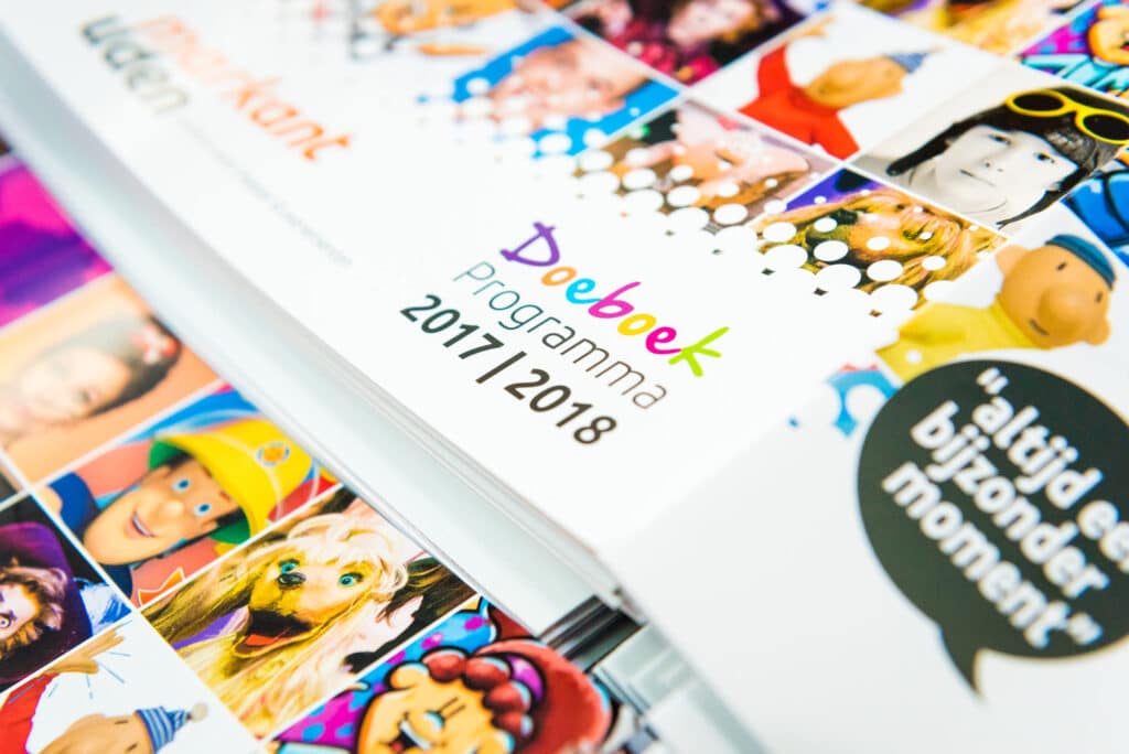 Een close-up van een stapel programmaboeken met de titel "Doeboek Programma 2017/2018" in kleurrijke letters. De kaft is bedekt met verschillende foto's van personages en illustraties. Rechts is een tekstballon met de tekst "altijd een bijzonder moment" te zien.
