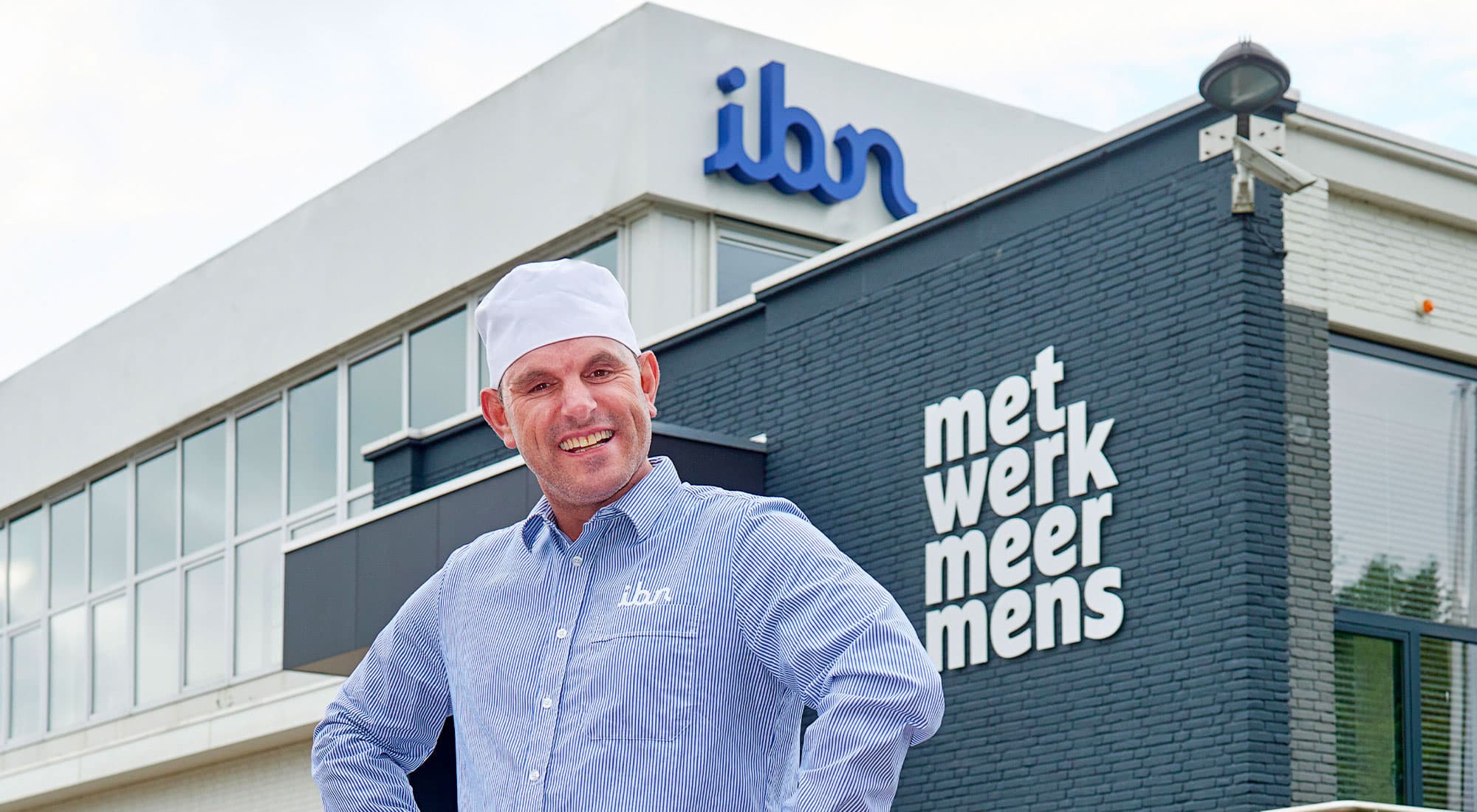 Een man in een lichtblauw gestreepte werkshirt en witte muts poseert lachend voor een modern gebouw met grote ramen. Op het gebouw staat de tekst "met werk meer mens" en het logo "ibn" is zichtbaar. Het is een zonnige dag.