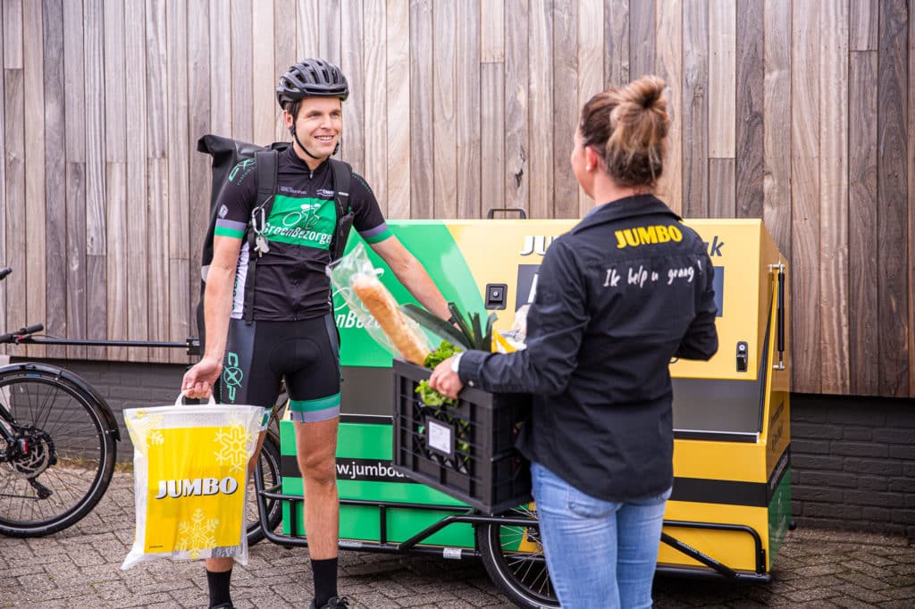 Een bezorger staat naast een bakfiets en geeft een Jumbo-boodschappentas aan een vrouw in een Jumbo-uniform. De vrouw houdt een krat vast met boodschappen, waaronder een stokbrood. De setting is buiten, voor een houten schutting.
