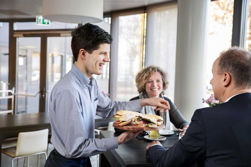 Een ober levert een bord met broodjes aan een tafel met twee gasten in een café-omgeving. De gasten, een lachende vrouw en een man in een pak, kijken blij naar de ober. Het interieur is modern met veel natuurlijk licht dat door grote ramen binnenkomt.
