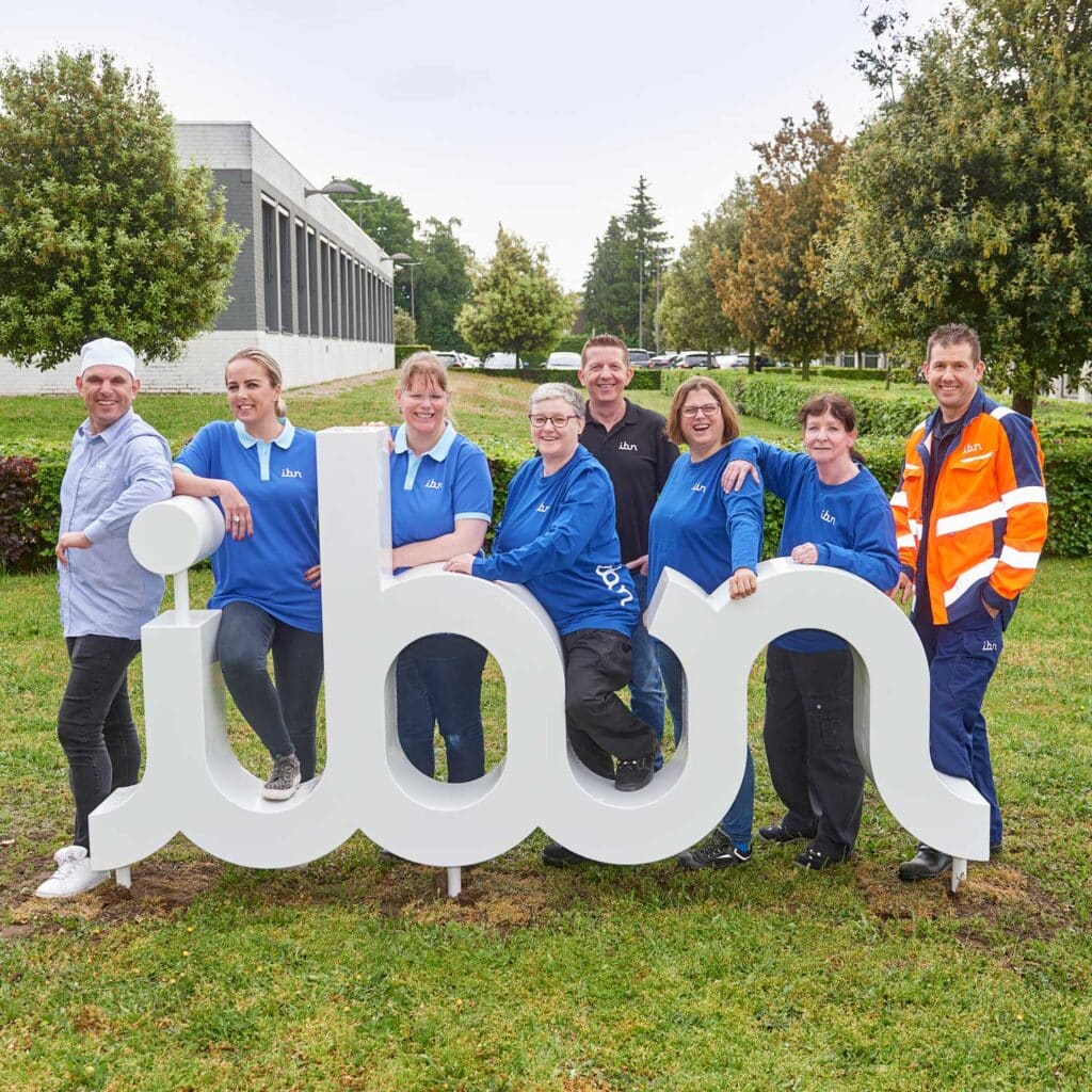 Een groep mensen poseert buiten bij een groot wit "ibn" logo. Ze dragen verschillende soorten werkkleding, waaronder blauwe shirts en een oranje veiligheidsjas. Op de achtergrond zijn groen gras, bomen en een gebouw te zien. Ze lachen en stralen een gevoel van saamhorigheid uit.
