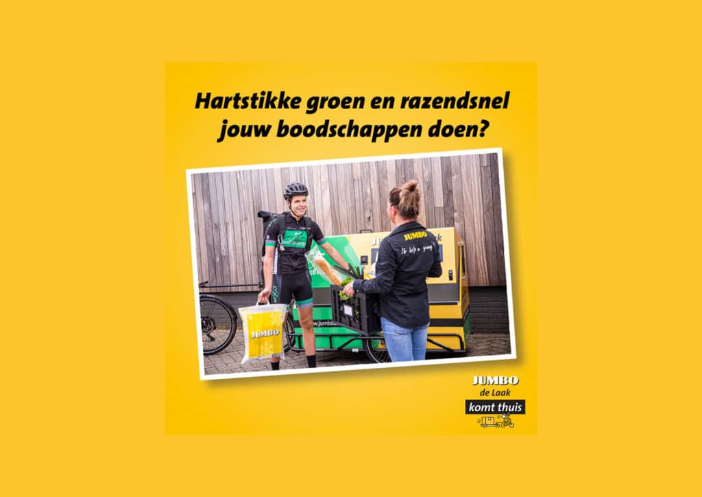 Een bezorger op een bakfiets overhandigt een tas met boodschappen aan een vrouw in een Jumbo-uniform. Op de achtergrond is een houten schutting te zien. Boven de foto staat de tekst "Hartstikke groen en razendsnel jouw boodschappen doen?" op een gele achtergrond. Linksonder staat het logo van "Jumbo de Laak komt thuis.''