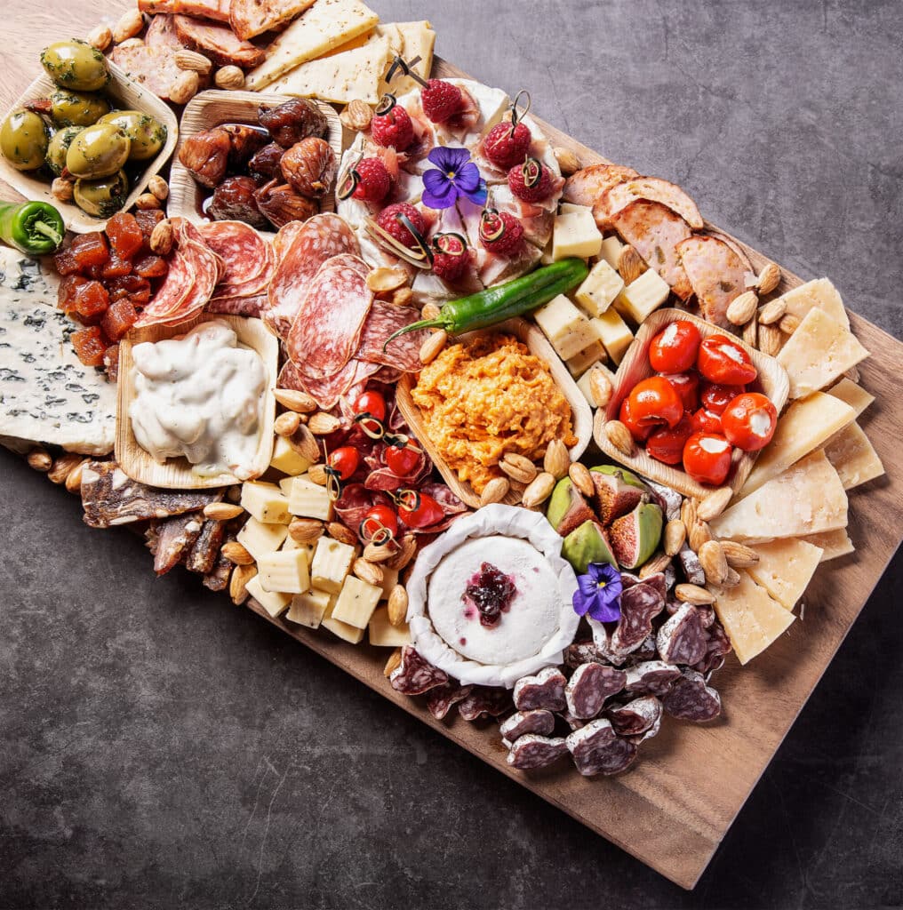 Een charcuterieplank vol met verschillende hapjes. Er liggen verschillende soorten kaas, vleeswaren, gevulde paprika's, olijven, noten, vijgen en dipsauzen op. Alles is kunstig gerangschikt op een houten plank, klaar om te serveren als luxe borrelhapje. Er zijn ook eetbare bloemen ter decoratie.