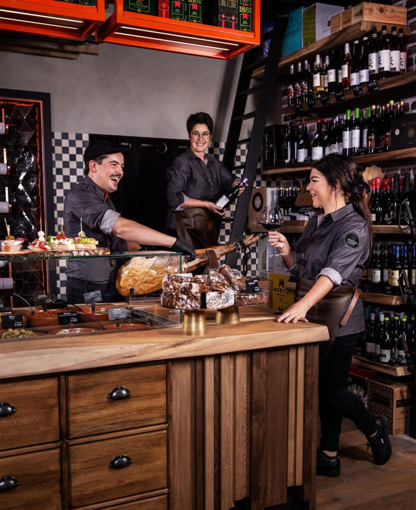 Drie medewerkers in een gezellige delicatessenwinkel lachen en werken samen. Een man snijdt ham terwijl een vrouw een glas wijn vasthoudt en een andere vrouw een fles wijn uit de rekken pakt. De winkel heeft een warme uitstraling met houten meubels, een uitgebreid wijnassortiment en een vitrine vol hapjes.