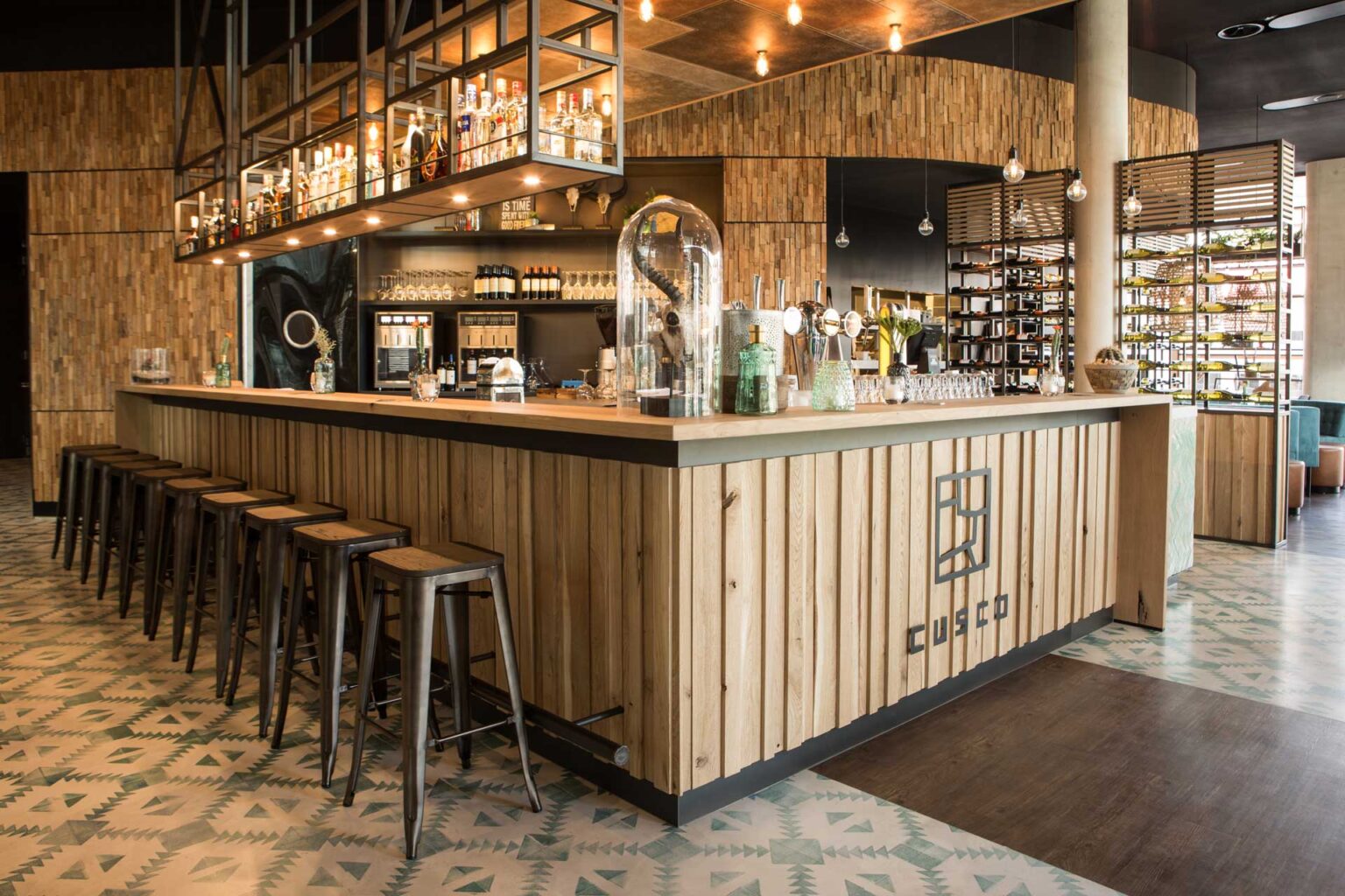 Een stijlvolle bar met een houten ontwerp en een reeks hoge metalen krukken. Boven de bar hangt een rek met flessen en glazen. De vloer heeft een patroon en er hangt warme verlichting. De ruimte heeft een moderne, uitnodigende sfeer met veel aandacht voor detail in de inrichting.
