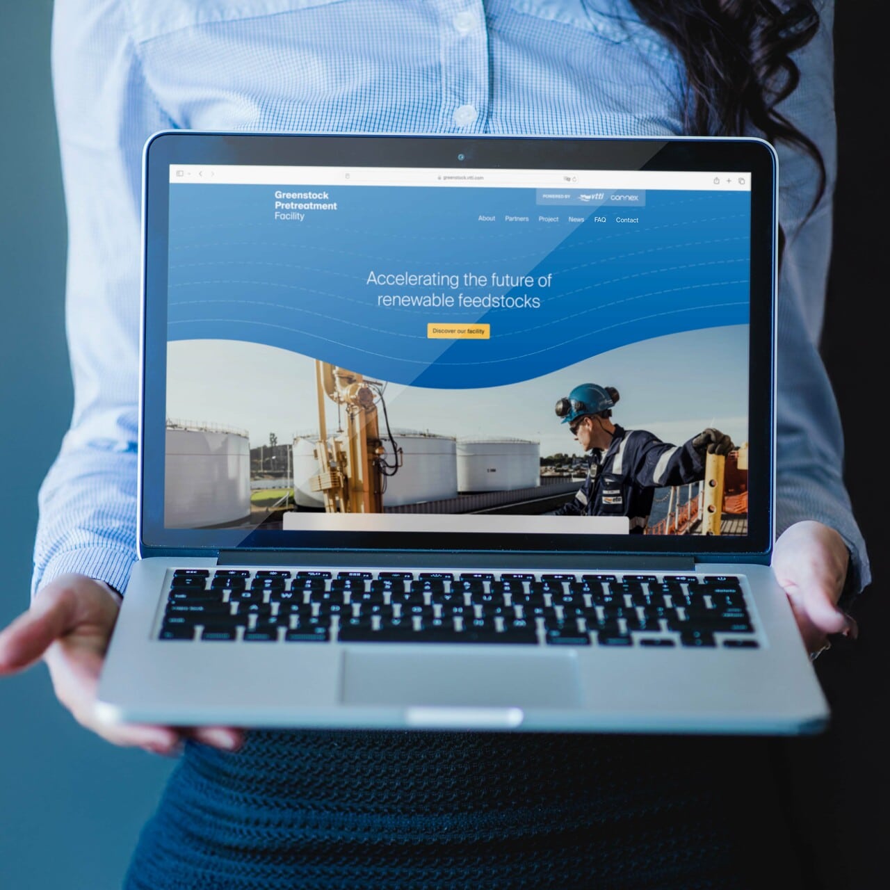 Een persoon in een blauw-wit gestreept overhemd houdt een open laptop vast, waarop een website wordt weergegeven. De website toont de titel "Greenstock Pretreatment Facility" met de slogan "Accelerating the future of renewable feedstocks". Op het scherm is een afbeelding te zien van een industrieel terrein met opslagtanks en een werknemer in een veiligheidshelm en werkpak.