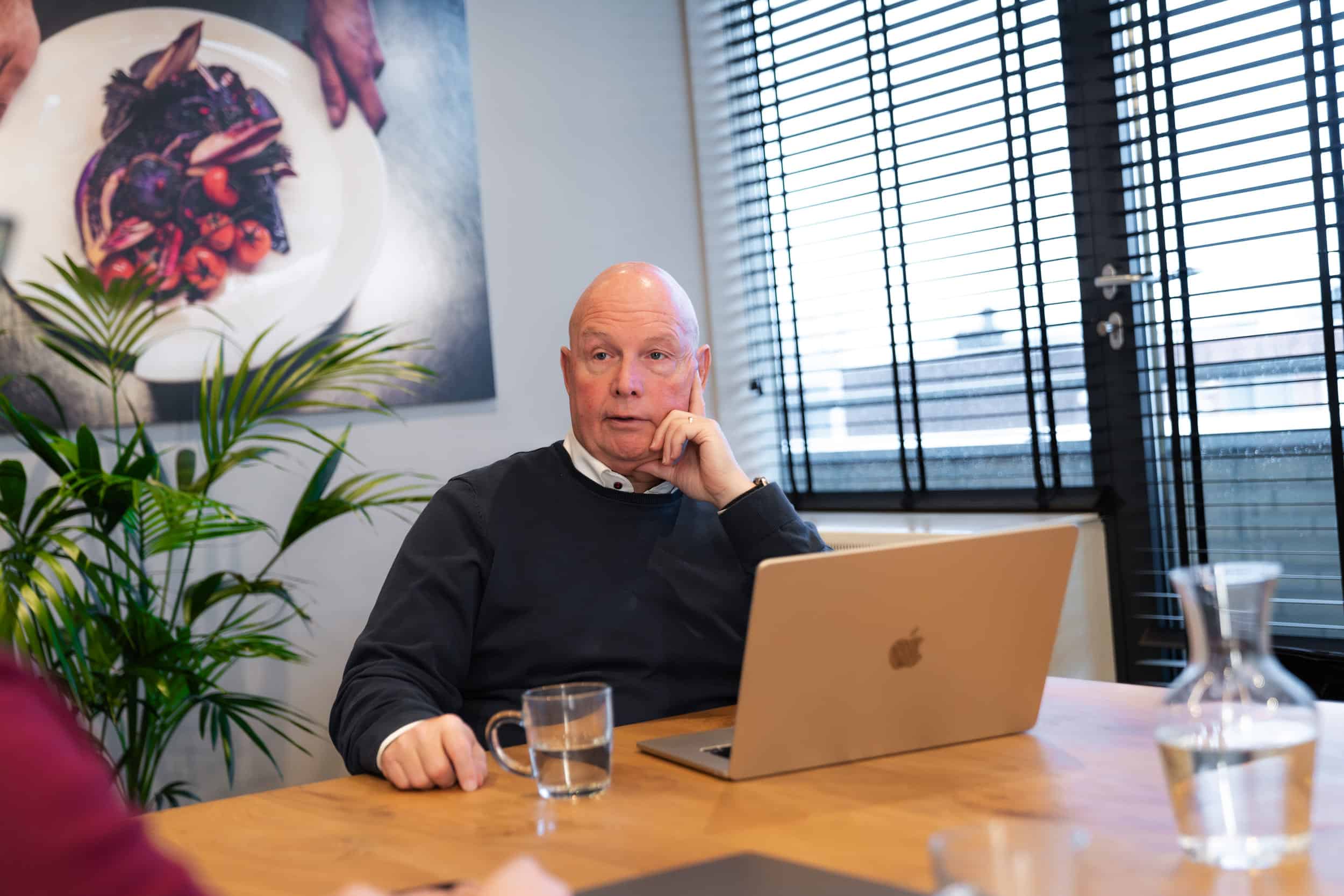 Een oudere man zit aan een vergadertafel met een opengeklapte MacBook en een glas water voor zich. Hij draagt een donkerblauwe trui met een wit overhemd en kijkt geconcentreerd richting iemand buiten beeld. Op de achtergrond zijn grote ramen met jaloezieën, een groene kamerplant en een grote foto van een culinair gerecht aan de muur zichtbaar. De sfeer is zakelijk en professioneel.