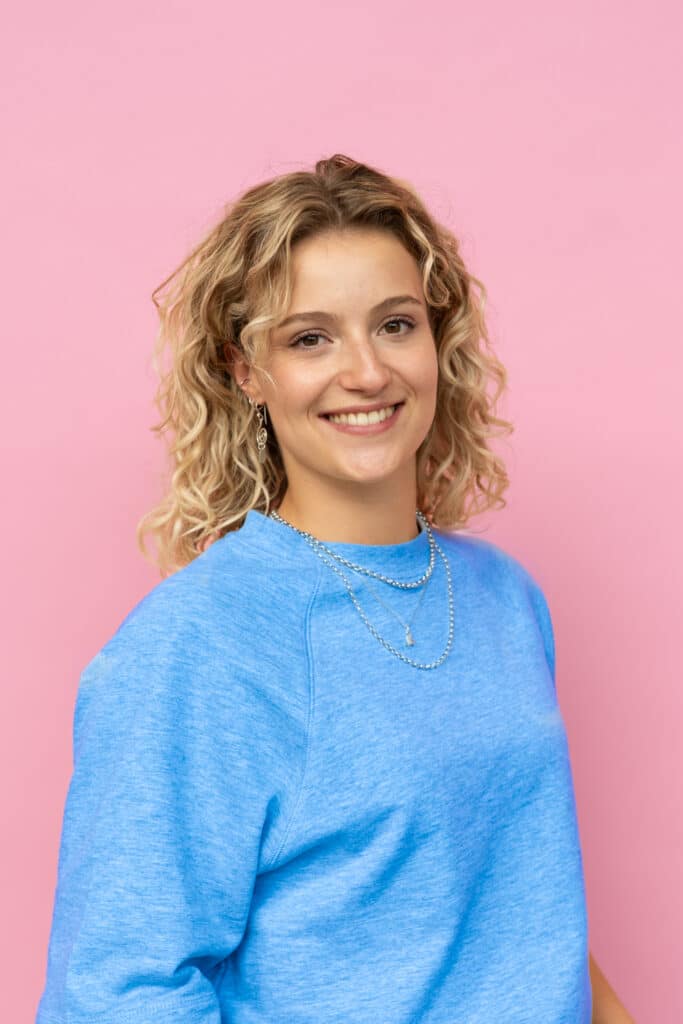Een jonge vrouw met blond, krullend haar glimlacht naar de camera. Ze draagt een blauwe trui, een lichte spijkerbroek en sieraden zoals kettingen, armbanden en oorbellen. Ze staat voor een roze achtergrond met haar handen nonchalant in haar broekzakken.