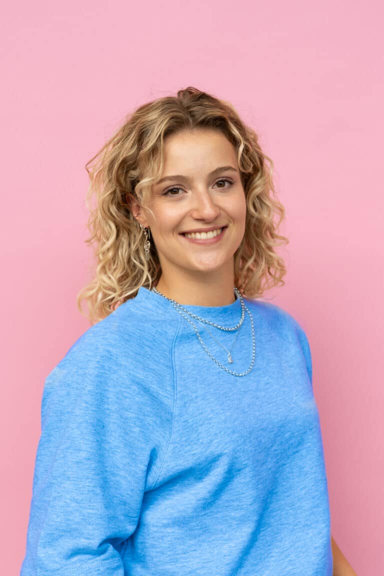Een jonge vrouw met blond, krullend haar glimlacht naar de camera. Ze draagt een blauwe trui, een lichte spijkerbroek en sieraden zoals kettingen, armbanden en oorbellen. Ze staat voor een roze achtergrond met haar handen nonchalant in haar broekzakken.