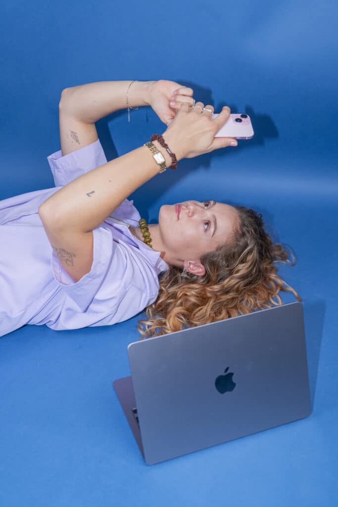 Vrouw ligt op een blauwe ondergrond met mobiel in haar hand en laptop naast haar.